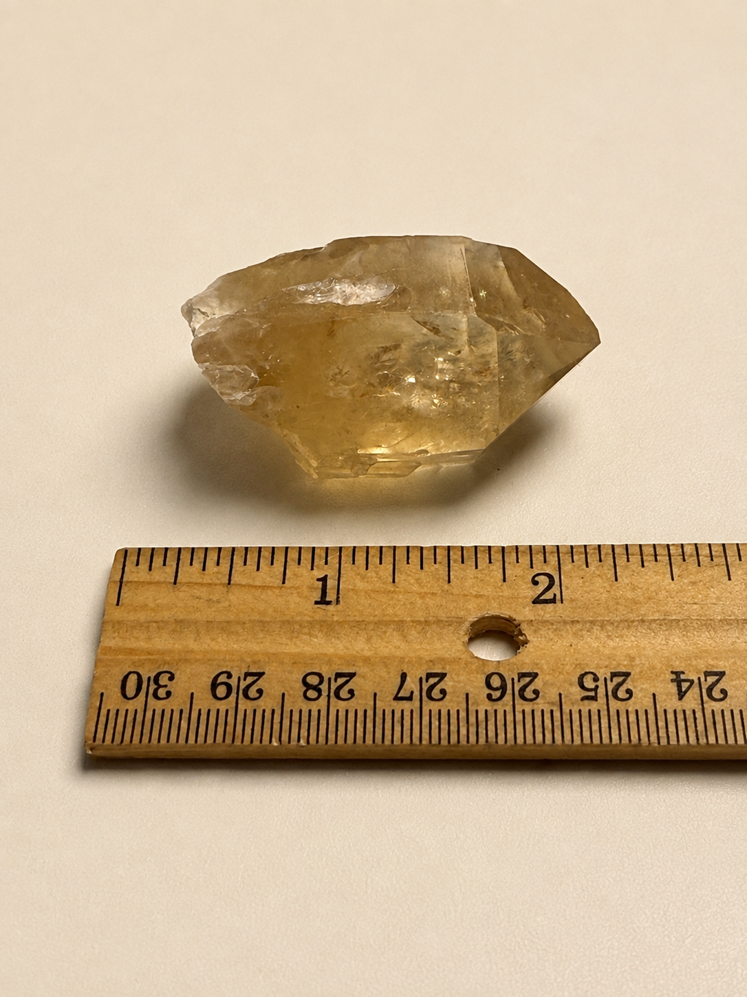 A rough, translucent yellow crystal specimen shown above a wooden ruler for scale on a plain light beige background.