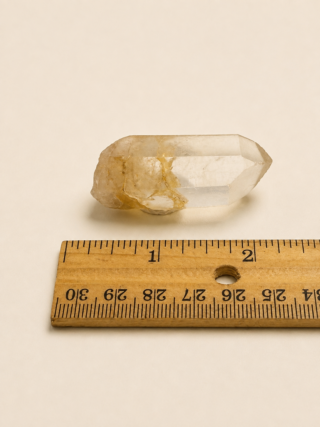 A small clear quartz crystal point with yellowish-beige inclusions, shown above a wooden ruler on a plain off-white background to indicate size.