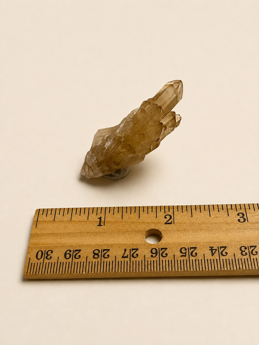 A small translucent yellowish crystal cluster placed above a wooden ruler on a plain off-white background, likely shown to indicate size.