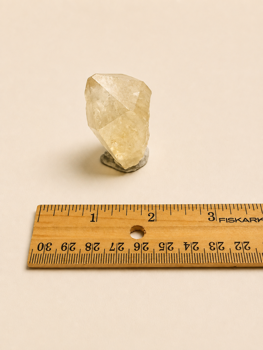 A small pale yellow crystal cluster on a dark stand, shown above a wooden ruler for scale on a warm off-white background.