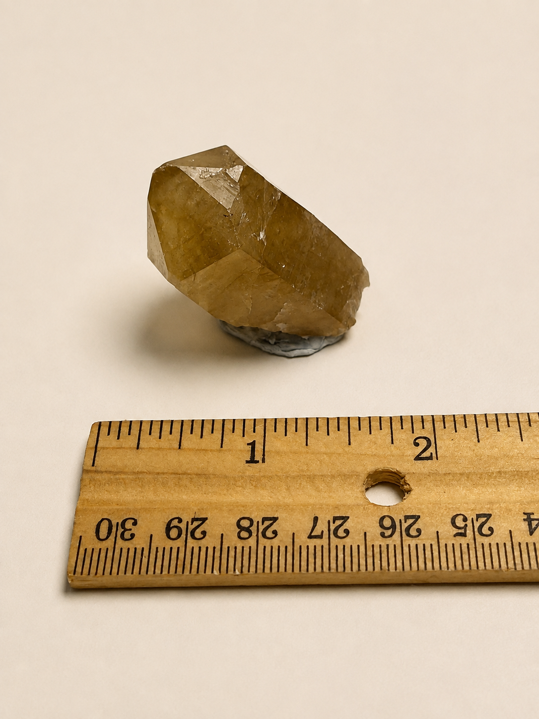 A faceted natural crystal stone with a warm golden-brown hue, displayed on a light beige background with a wooden ruler below it for scale.