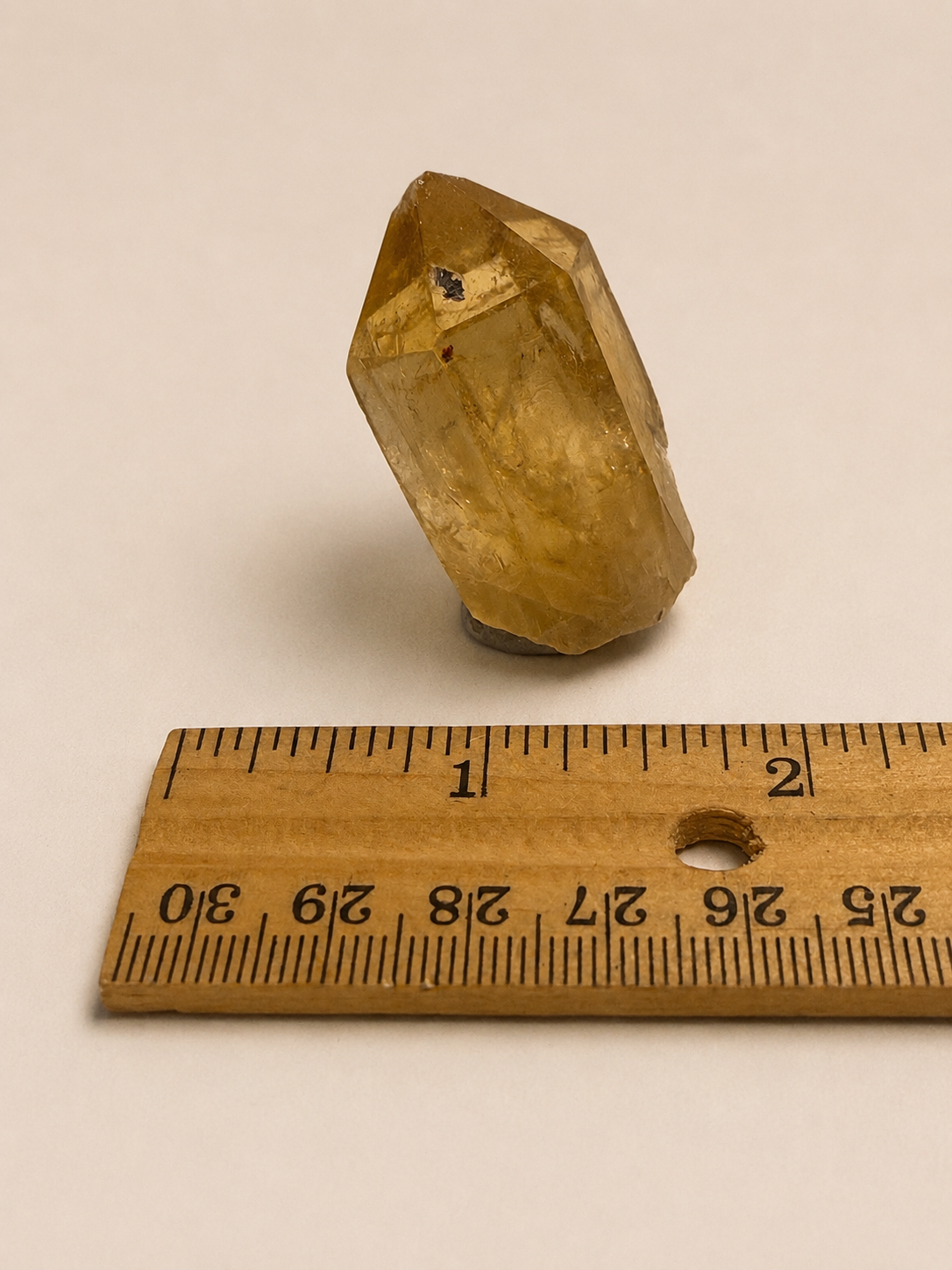 A small natural yellow crystal or quartz point standing upright on a plain light background, with a wooden ruler placed below it for scale.