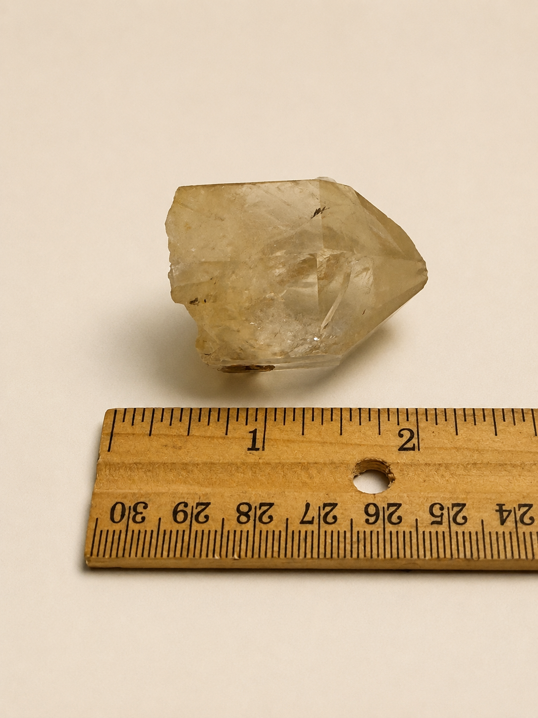 A small translucent pale yellow crystal or stone placed above a wooden ruler on a plain off-white background, with the ruler showing measurements in centimeters.