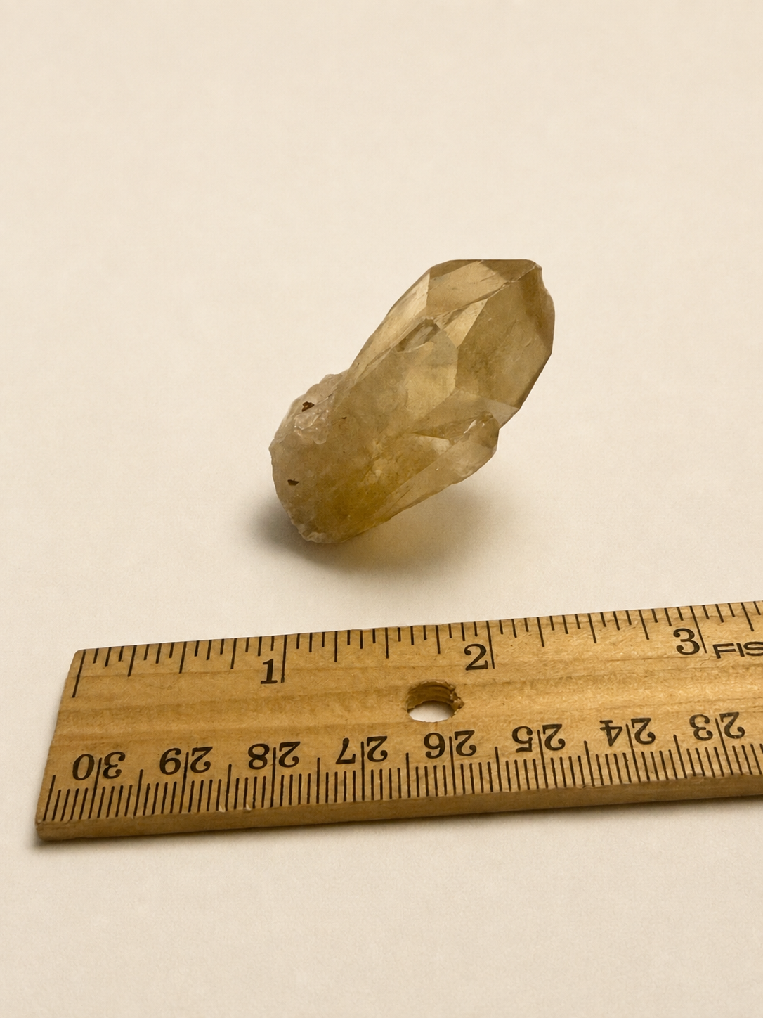 A small translucent pale yellow crystal specimen placed above a wooden ruler on a plain off-white background, likely shown for size reference.