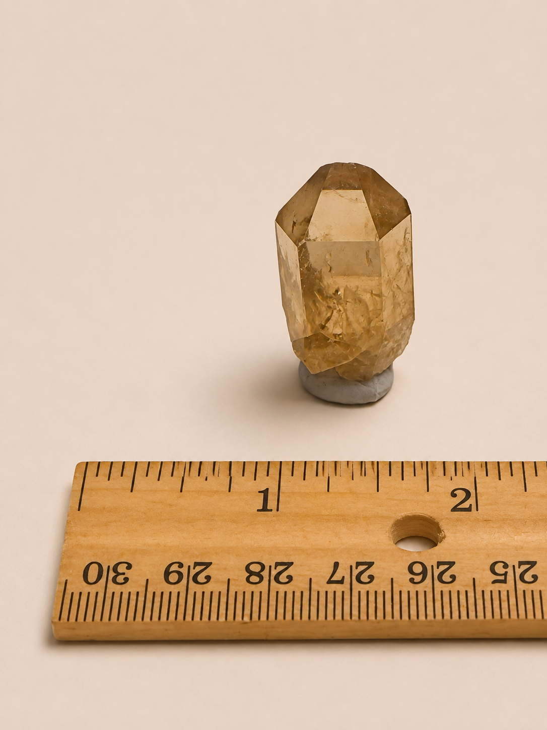 A small faceted translucent yellow crystal or gemstone mounted on a gray base, shown above a wooden ruler on a plain warm beige background.