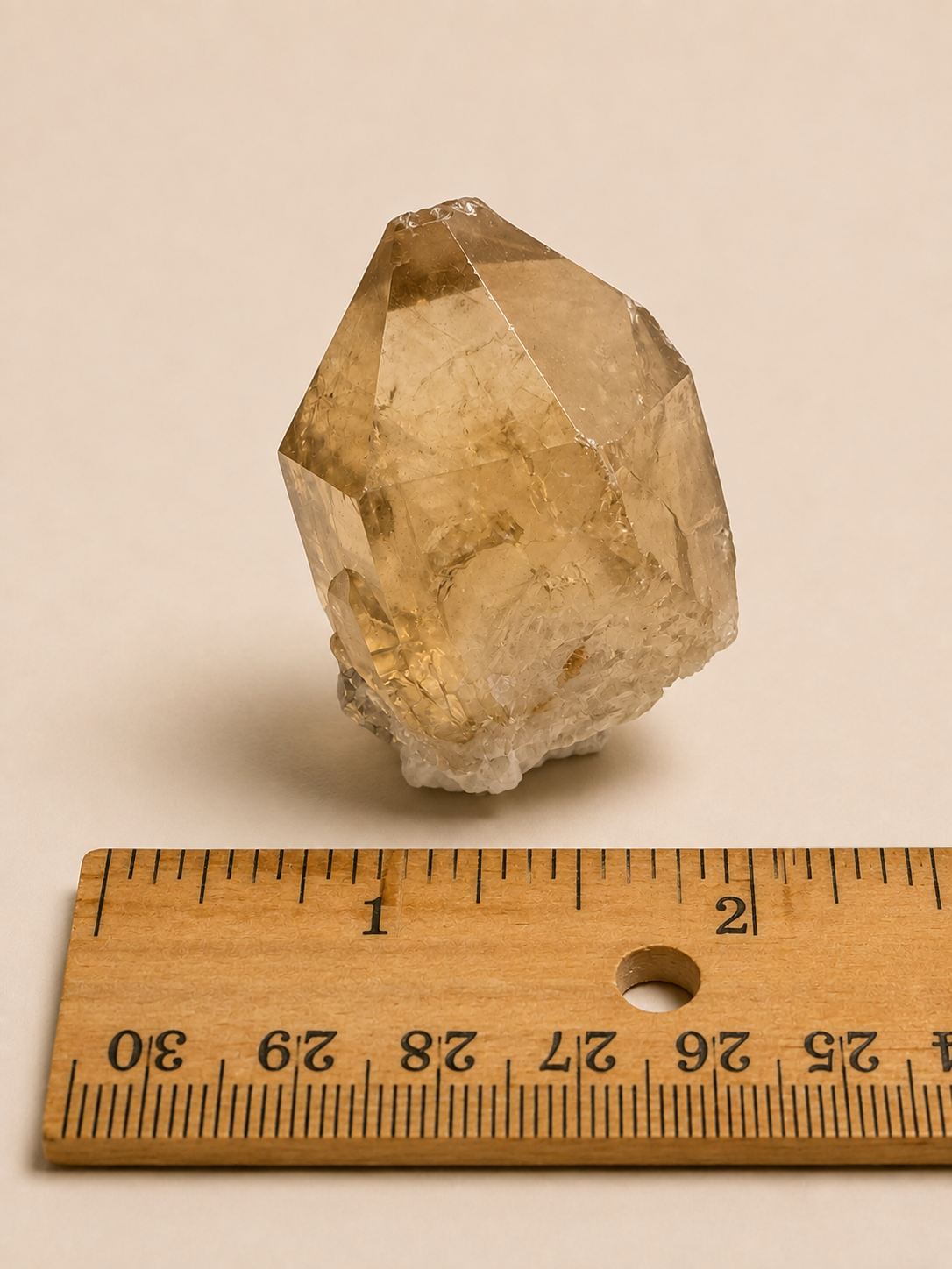 A natural clear quartz crystal point with warm golden-brown and translucent tones, shown next to a wooden ruler for scale on a plain beige background.