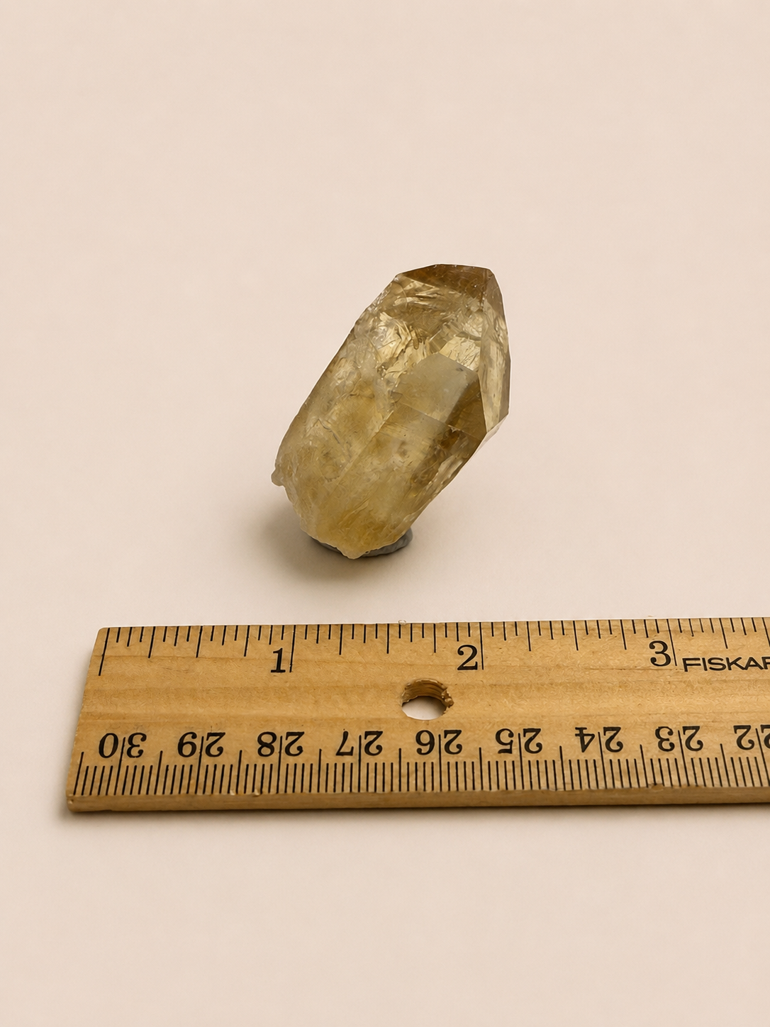 A natural faceted crystal stone with a warm yellow-brown hue, shown next to a wooden ruler for scale on a plain light beige background.