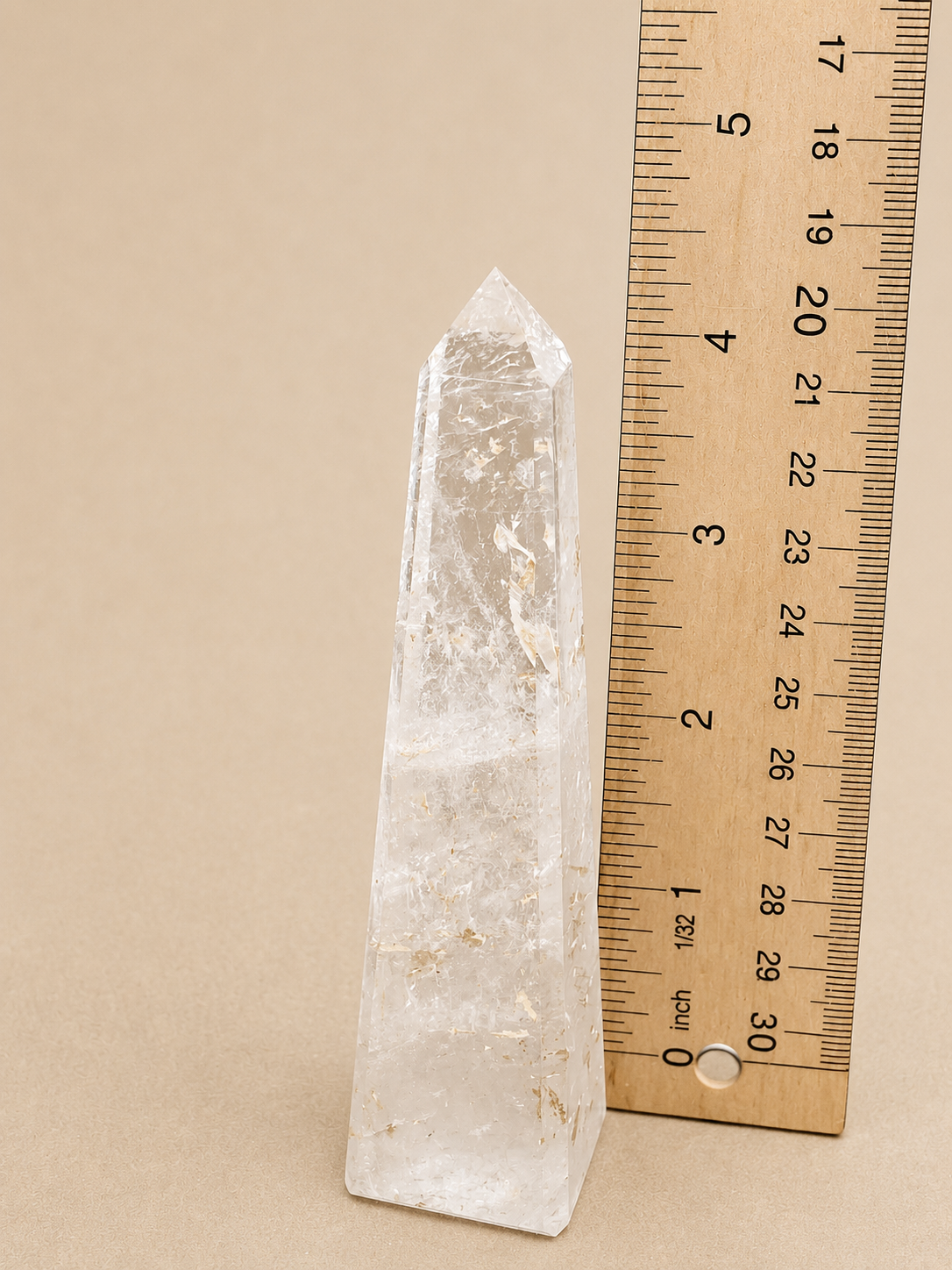 A clear quartz crystal point tower with natural inclusions, shown next to a wooden ruler for scale on a solid beige background.
