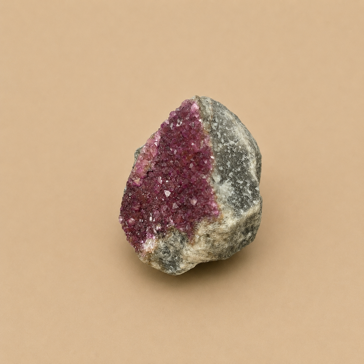A natural mineral specimen featuring a prominent magenta-purple crystal cluster on the left side of a gray and white textured rock matrix, set against a solid beige background.