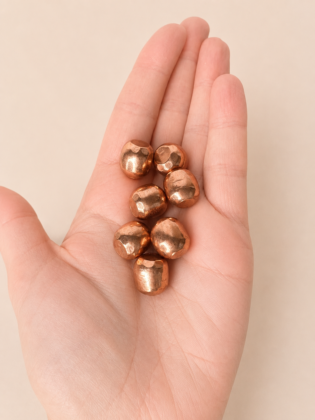 A close-up of a person's hand holding several small, metallic copper-colored beads against a soft beige background.