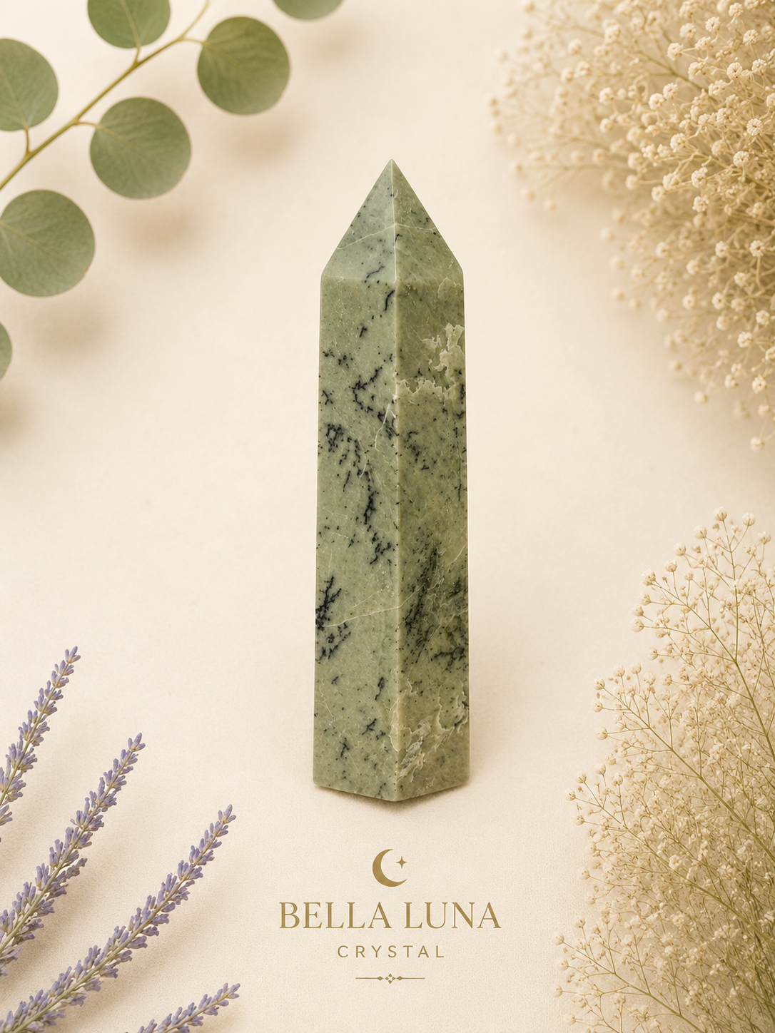 A polished green crystal obelisk with natural marbling, displayed on a warm cream background with botanical props and the Bella Luna Crystal logo at the bottom.