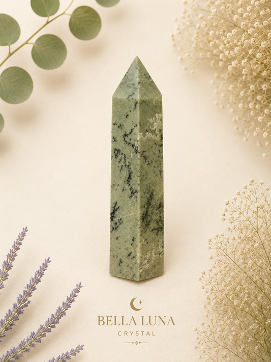 A polished green crystal obelisk with natural marbling, displayed on a warm cream background with botanical props and the Bella Luna Crystal logo at the bottom.