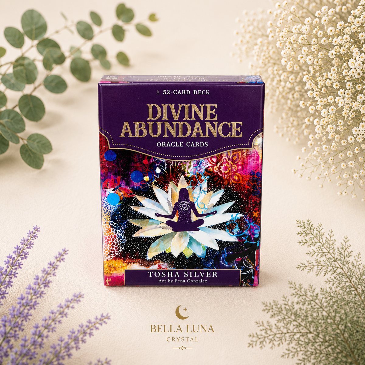 Divine Abundance Oracle Cards deck box by Tosha Silver, styled on a light beige surface with botanical elements like eucalyptus, baby's breath, and lavender. The box features a purple top section with gold lettering and a colorful, abstract lotus design on the front.