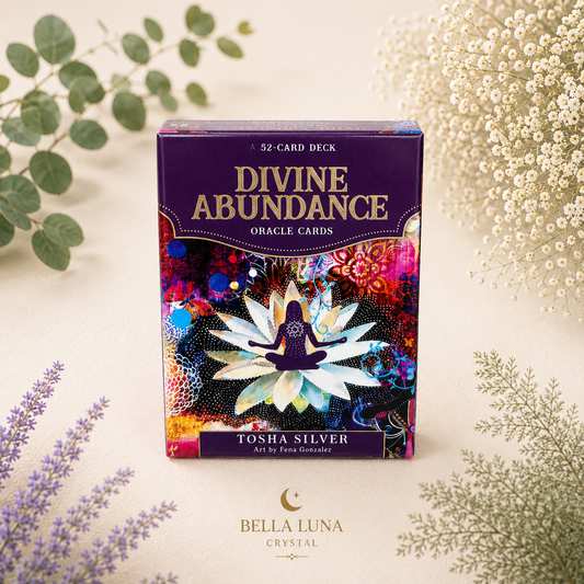 Divine Abundance Oracle Cards deck box by Tosha Silver, styled on a light beige surface with botanical elements like eucalyptus, baby's breath, and lavender. The box features a purple top section with gold lettering and a colorful, abstract lotus design on the front.