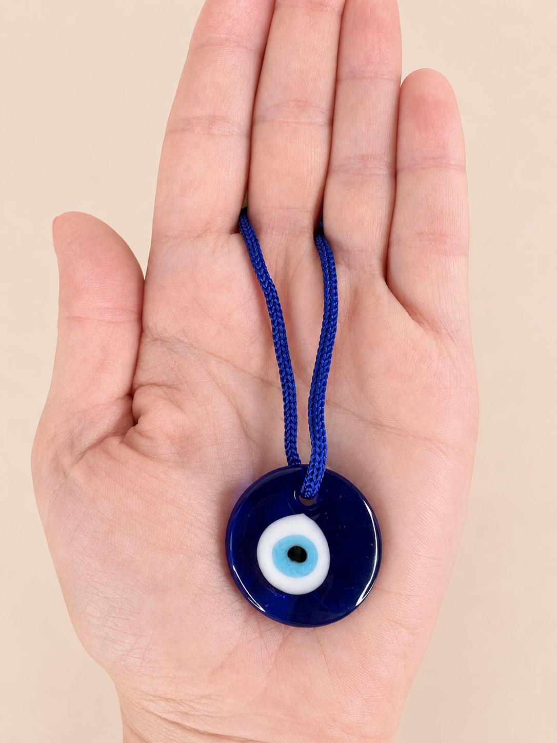 A blue glass evil eye pendant necklace with a blue cord, held in the palm of a hand against a solid beige background.