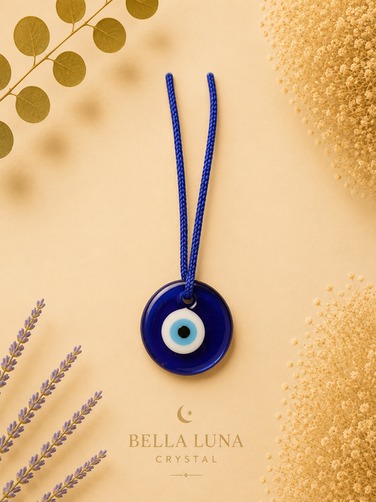 A blue glass evil eye pendant necklace with a blue braided cord, displayed on a warm beige background with decorative dried flowers and leaves. The brand name 'BELLA LUNA CRYSTAL' is printed at the bottom.