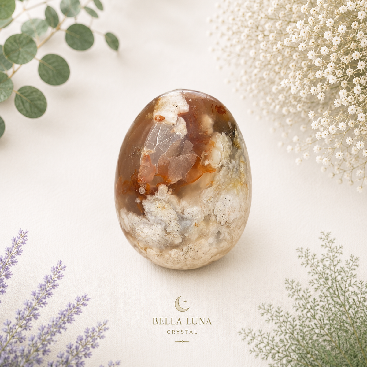 A polished oval crystal stone with natural brown, amber, white, and gray inclusions, displayed on a light cream surface with decorative green and white flowers. The brand name Bella Luna Crystal is printed at the bottom.
