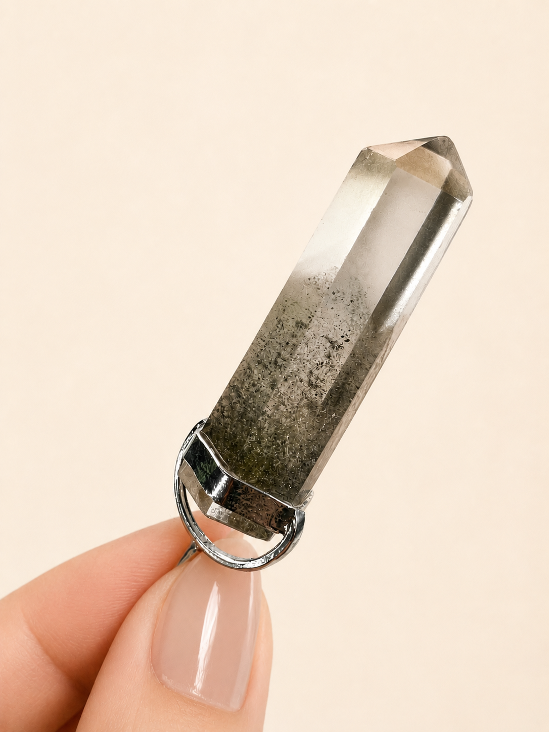 A smoky quartz crystal point pendant with a silver metal ring setting, held between fingers against a solid light beige background.