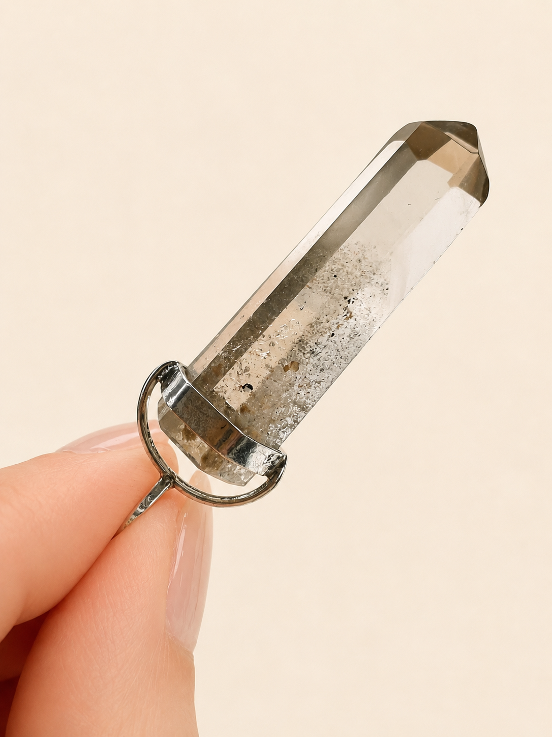 A clear quartz crystal point pendant in a silver bezel setting, held by a person's fingers against a solid light beige background.