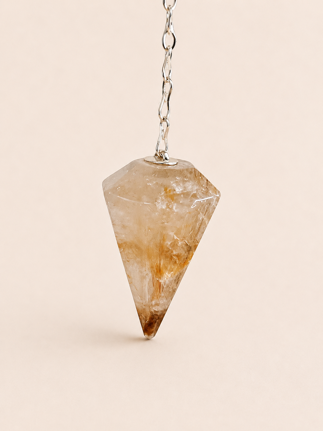 A faceted crystal pendulum pendant with a silver chain, suspended against a solid light beige background.