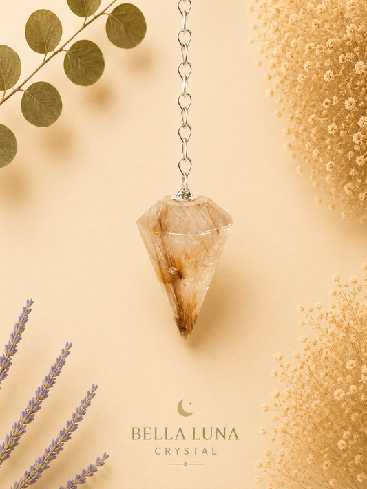 A faceted golden-rutile-pendulum clear crystal pendant necklace with a silver chain, hanging against a warm beige background with dried botanical props and the Bella Luna Crystal logo at the bottom.