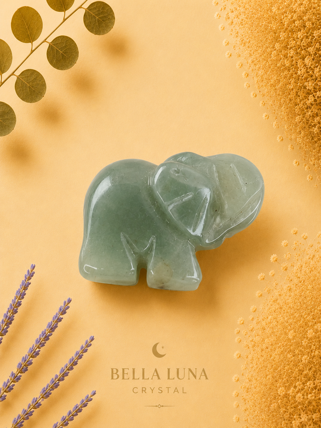 A carved pale green crystal elephant figurine centered on a warm yellow background, with decorative dried lavender stems, gold leaves, and textured gold elements around the edges. The brand name Bella Luna Crystal appears near the bottom.