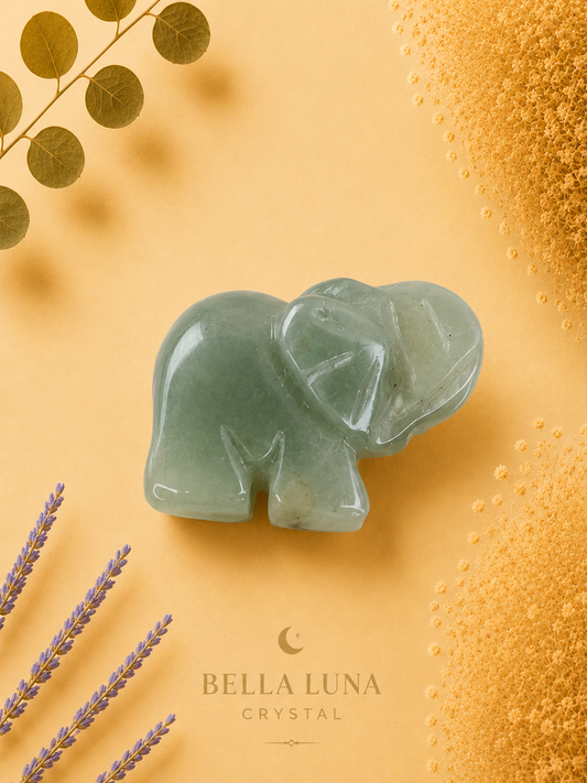 A carved pale green crystal elephant figurine centered on a warm yellow background, with decorative dried lavender stems, gold leaves, and textured gold elements around the edges. The brand name Bella Luna Crystal appears near the bottom.