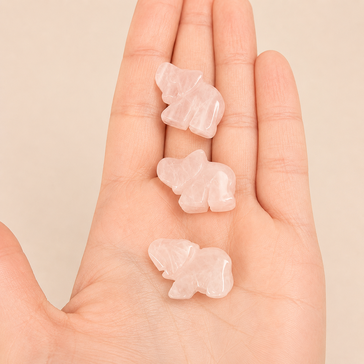 Three small pale pink carved elephant figurines arranged vertically on an open human hand against a soft beige background.