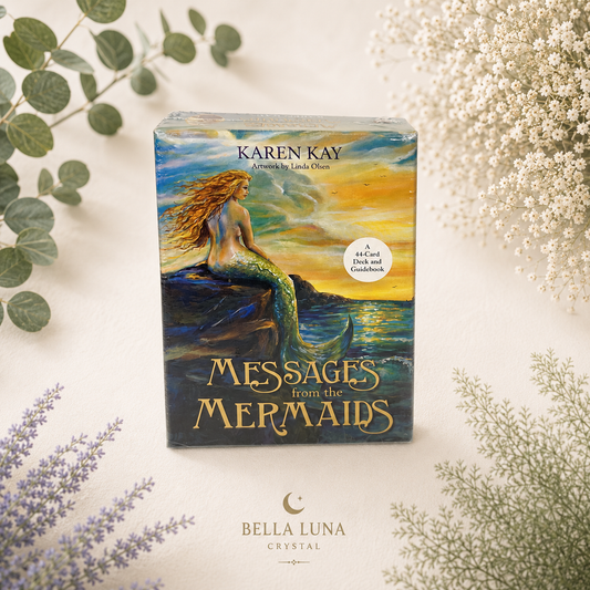 A boxed card deck titled "Messages from the Mermaids" by Karen Kay, placed on a light cream surface with decorative greenery and white flowers around it. The box art shows a mermaid sitting on rocks by a glowing sunset, and the Bella Luna Crystal logo appears below the box.