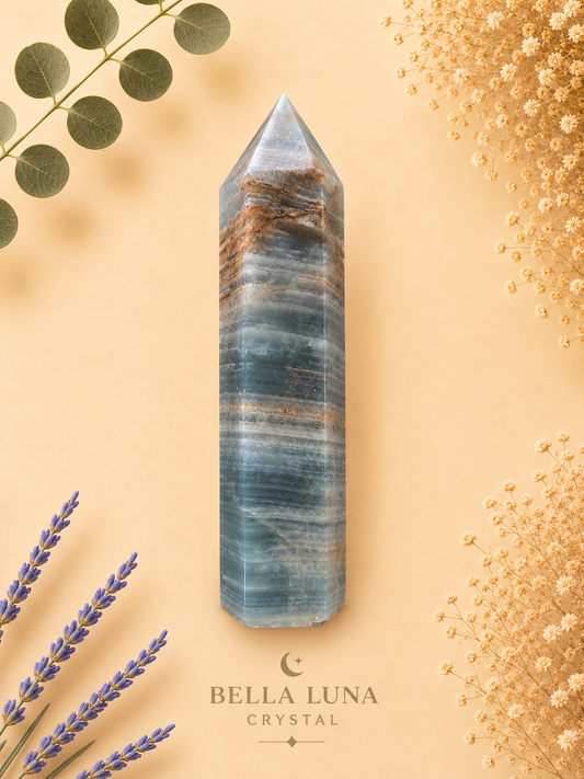 A tall blue-gray crystal point with natural brown and white banding, displayed on a warm beige background with decorative dried flowers, lavender, and eucalyptus. The brand name 'BELLA LUNA CRYSTAL' is printed at the bottom.