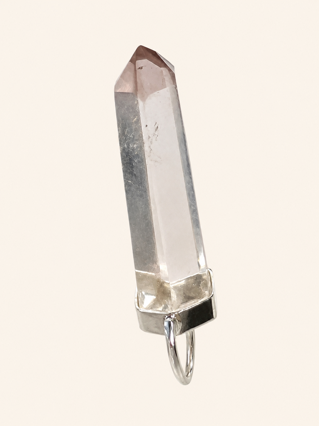 A clear quartz crystal point pendant with a silver metal cap and hanging loop, shown against a solid pale cream background.