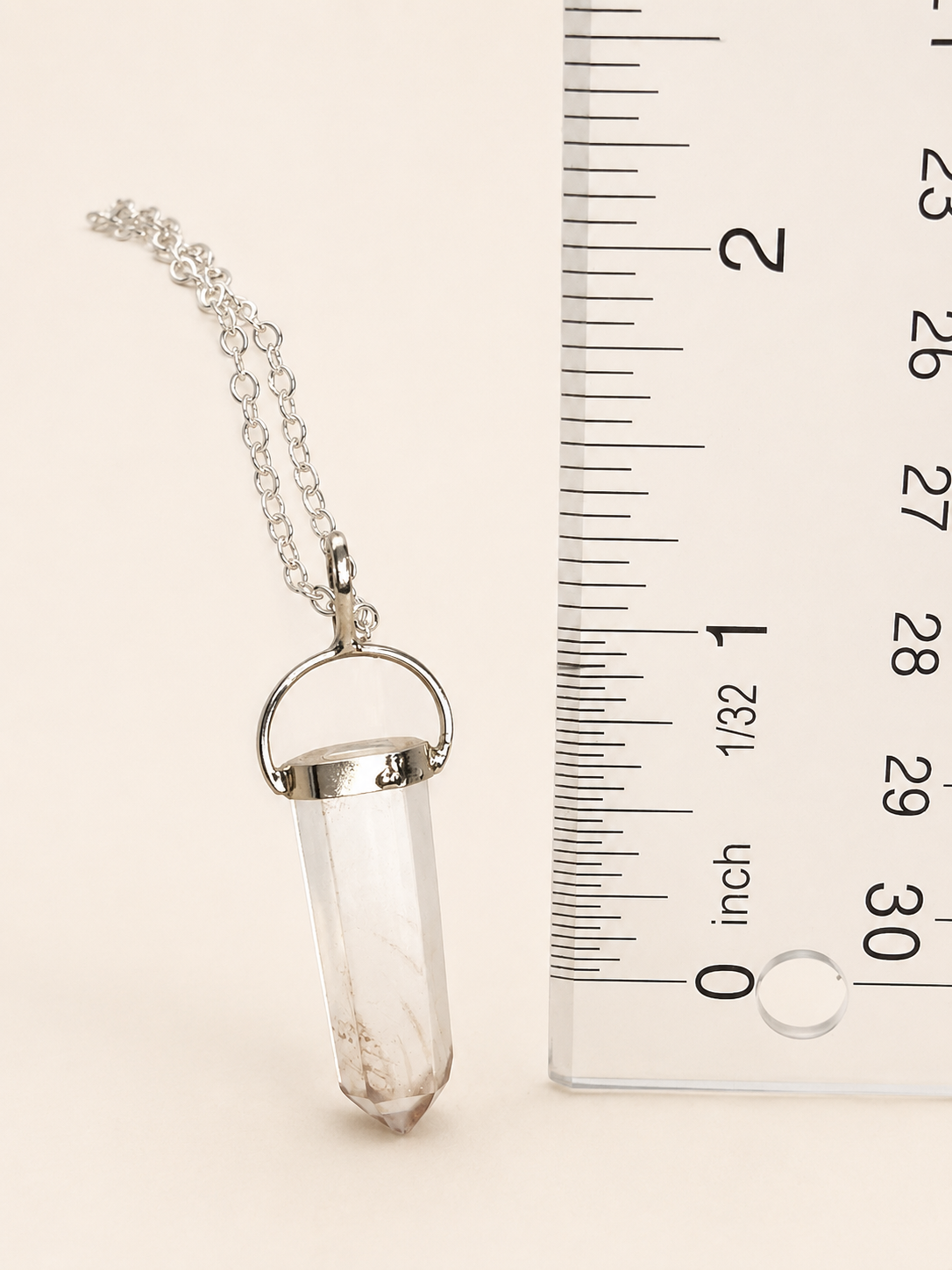 A silver-toned crystal pendant necklace on a delicate chain, shown next to a clear ruler for scale on a light beige background.