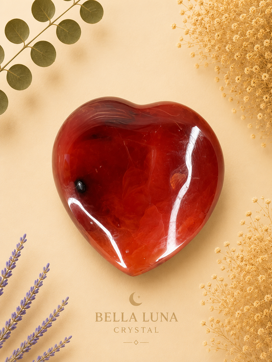 A polished red heart-shaped crystal stone with natural marbling, displayed on a warm beige background with decorative dried flowers and leaves. The brand name 'BELLA LUNA CRYSTAL' is printed at the bottom.