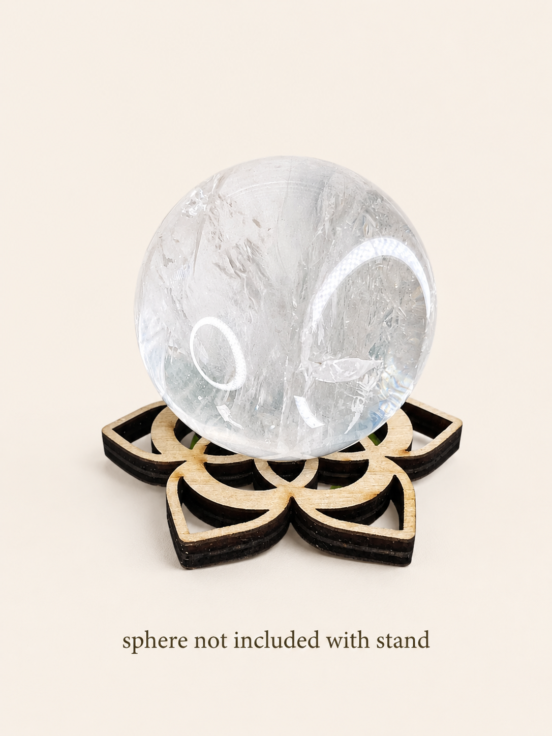 A clear quartz crystal sphere resting on a black and natural wood geometric stand, set against a solid beige background with the text 'sphere not included with stand' below.