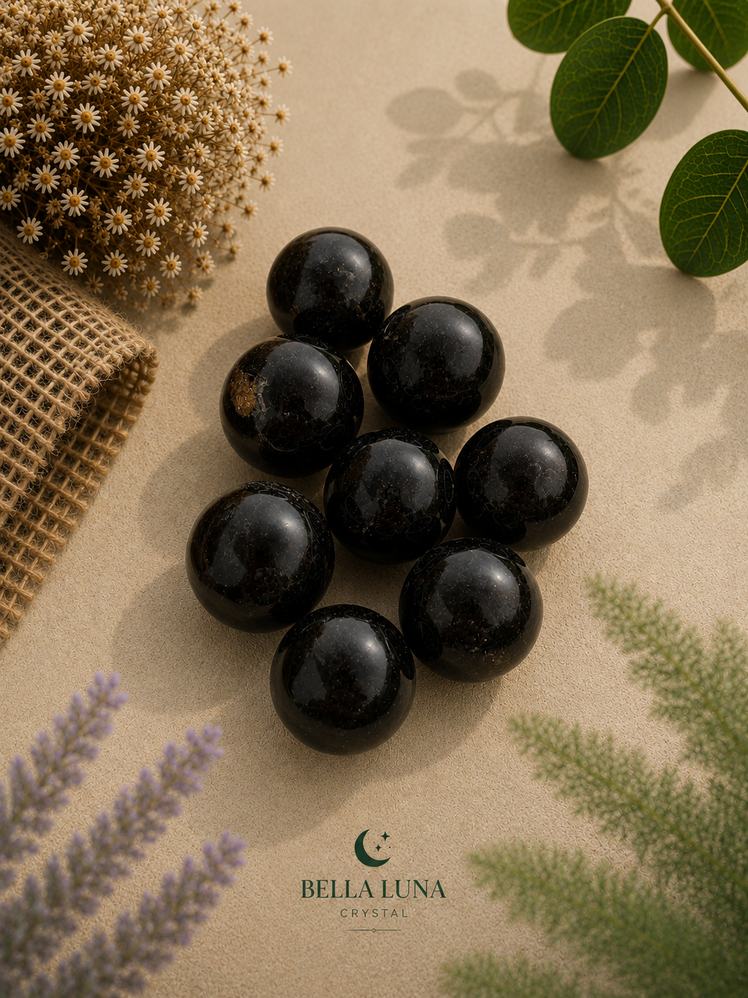 Nine polished black gemstone spheres arranged in a cluster on a textured beige surface, styled with dried flowers, green leaves, and burlap. The brand name 'BELLA LUNA CRYSTAL' is printed at the bottom.