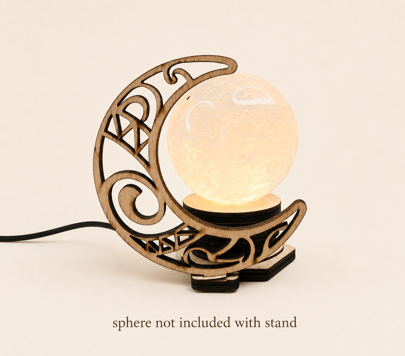 A crescent moon-shaped wooden lamp stand with intricate scrollwork, holding a glowing translucent sphere on a small black base. A black power cord extends from the left, and text at the bottom reads 'sphere not included with stand' on a warm cream background.