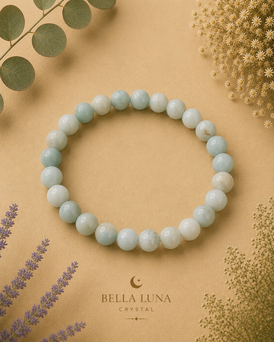 A beaded stretch bracelet made of light blue-green round stones, arranged in a circle on a warm beige background with dried botanicals and the Bella Luna Crystal logo printed below.