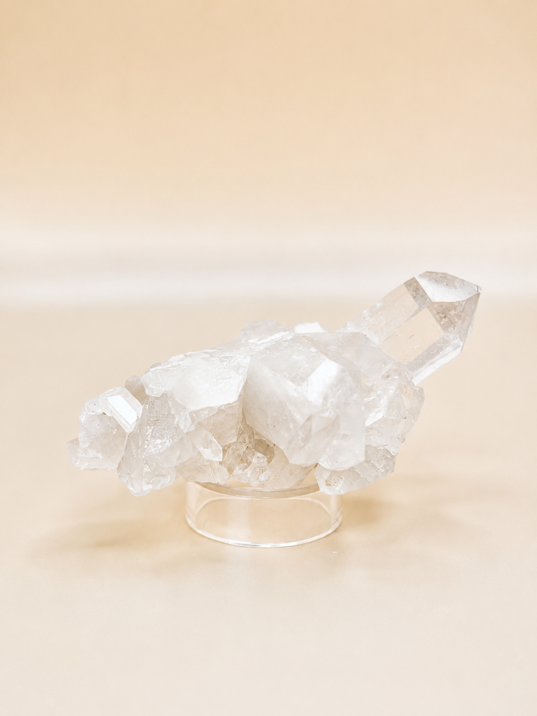 A clear quartz crystal cluster on a transparent acrylic display stand, set against a warm beige background.