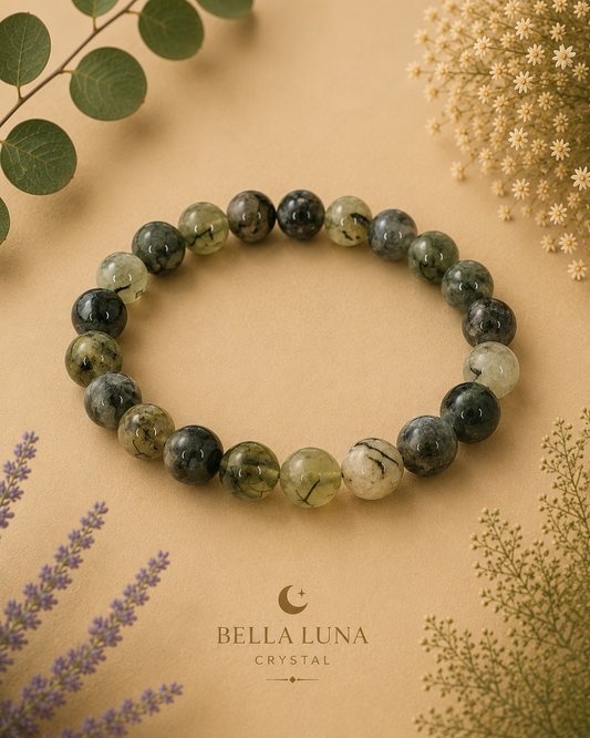 A beaded stretch bracelet made of polished green and black stone beads, displayed on a warm beige background with decorative dried flowers and leaves. The brand logo 'BELLA LUNA CRYSTAL' is printed below the bracelet.