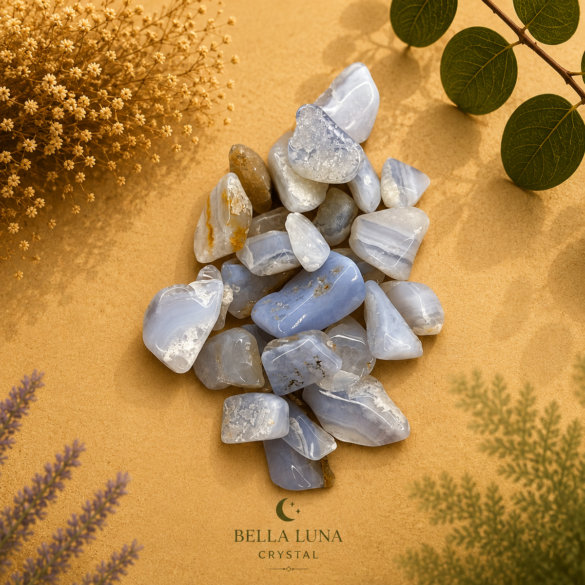 A pile of polished tumbled crystals in soft blue, gray, and white tones arranged on a warm tan surface with dried flowers and green leaves around them. The brand name BELLA LUNA CRYSTAL appears at the bottom.