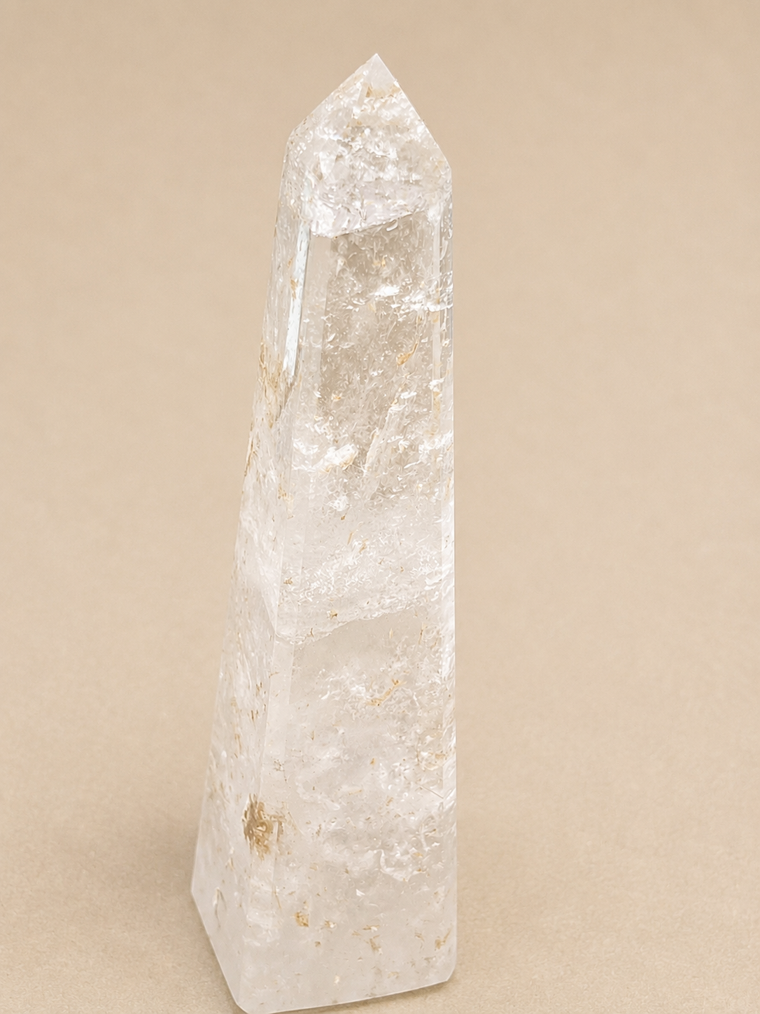 A natural clear quartz crystal tower with a pointed tip, standing upright on a solid beige background.