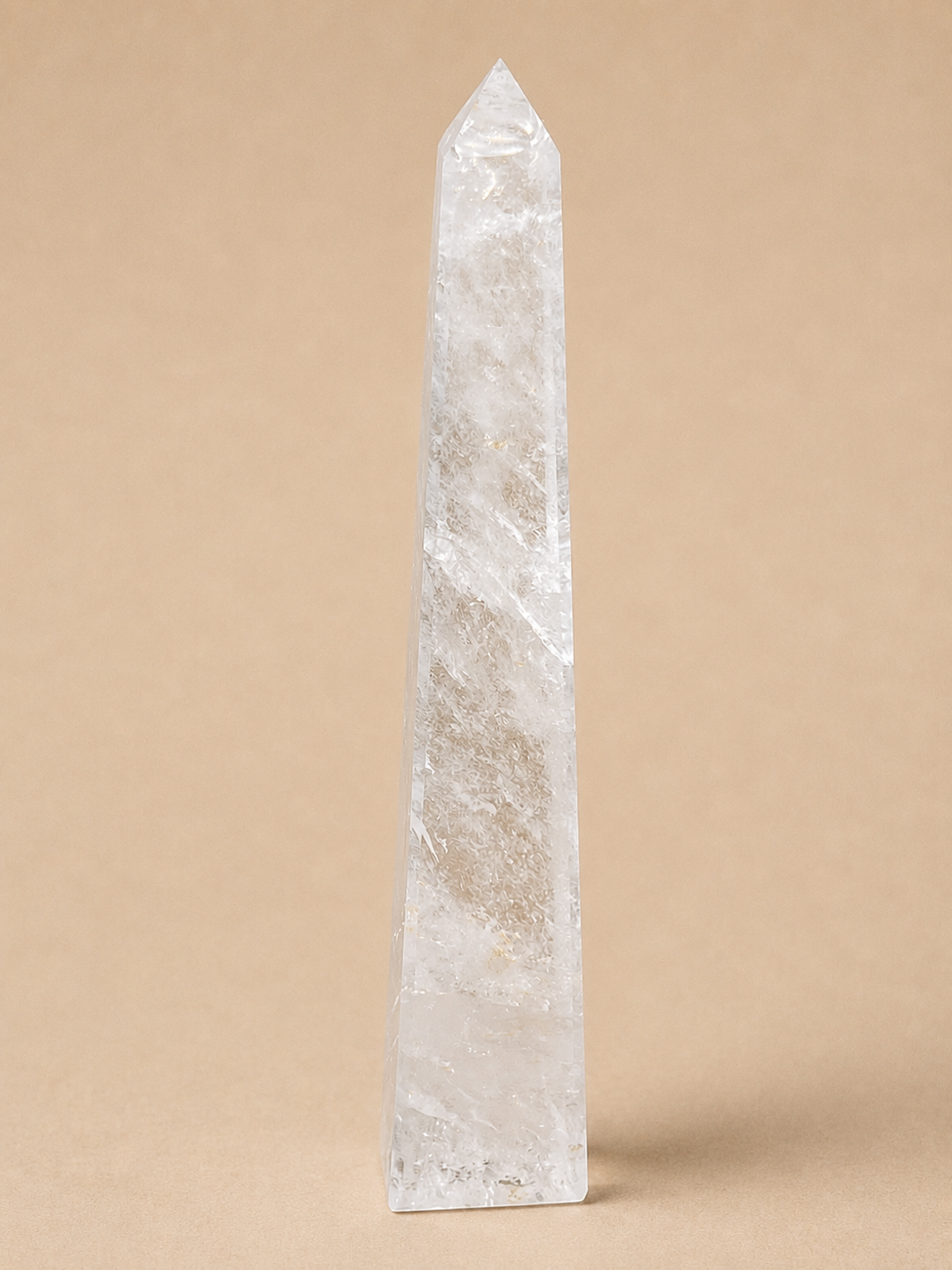 A clear quartz crystal obelisk with natural inclusions, standing upright on a solid beige background.