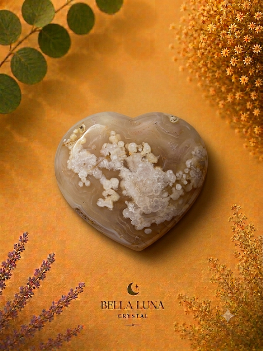 A heart-shaped crystal with white cloudy inclusions on a warm amber background, surrounded by dried flowers and green leaves. The brand name 'BELLA LUNA CRYSTAL' is printed at the bottom.