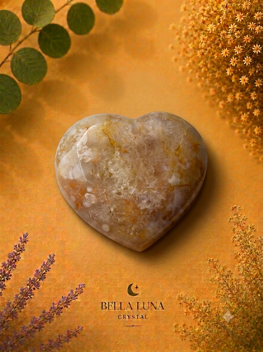 A polished heart-shaped crystal with natural beige, white, and golden-brown marbling, displayed on a textured golden-orange background with botanical elements and the Bella Luna Crystal brand logo.
