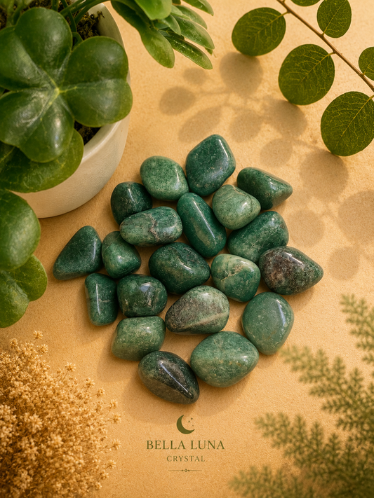 A pile of polished green tumbled stones arranged on a beige surface with decorative plants and dried flowers. The brand name Bella Luna Crystal appears at the bottom.