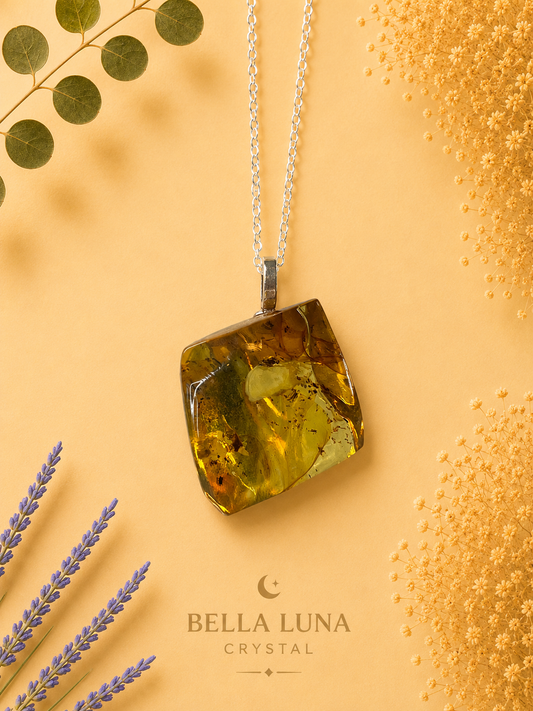 A square-cut yellow-green crystal pendant necklace with inclusions, suspended from a silver chain, displayed on a warm beige background with decorative dried lavender, eucalyptus leaves, and gold glitter. The brand name 'BELLA LUNA CRYSTAL' is printed at the bottom.