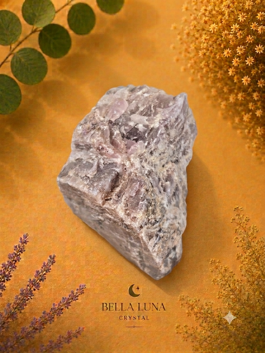 A rough, polished crystal stone with natural jagged edges and a mottled gray and white surface, displayed on a warm golden-brown textured background with botanical elements. The brand name 'BELLA LUNA CRYSTAL' is printed at the bottom.