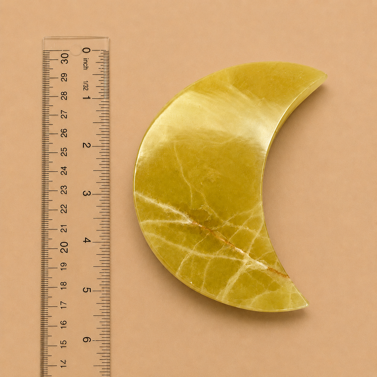 A polished crescent moon-shaped yellow stone pendant or brooch displayed next to a clear plastic ruler on a solid tan background.