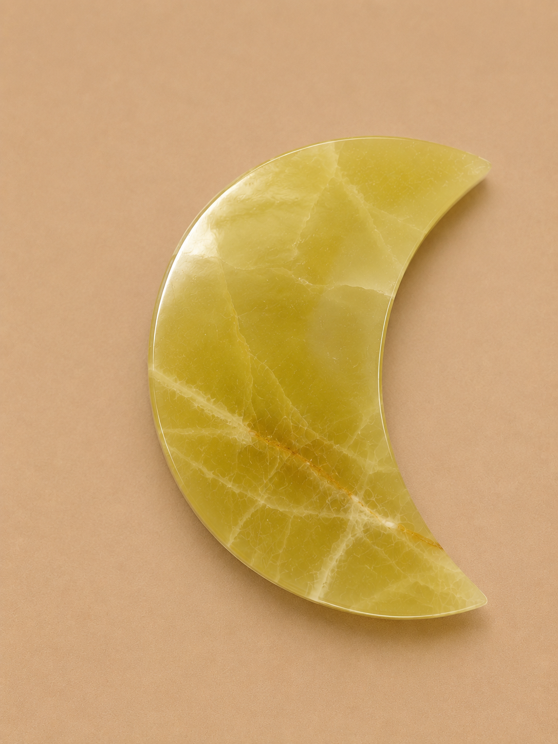 A crescent moon-shaped polished stone accessory in pale yellow with natural veining, displayed on a solid tan background.