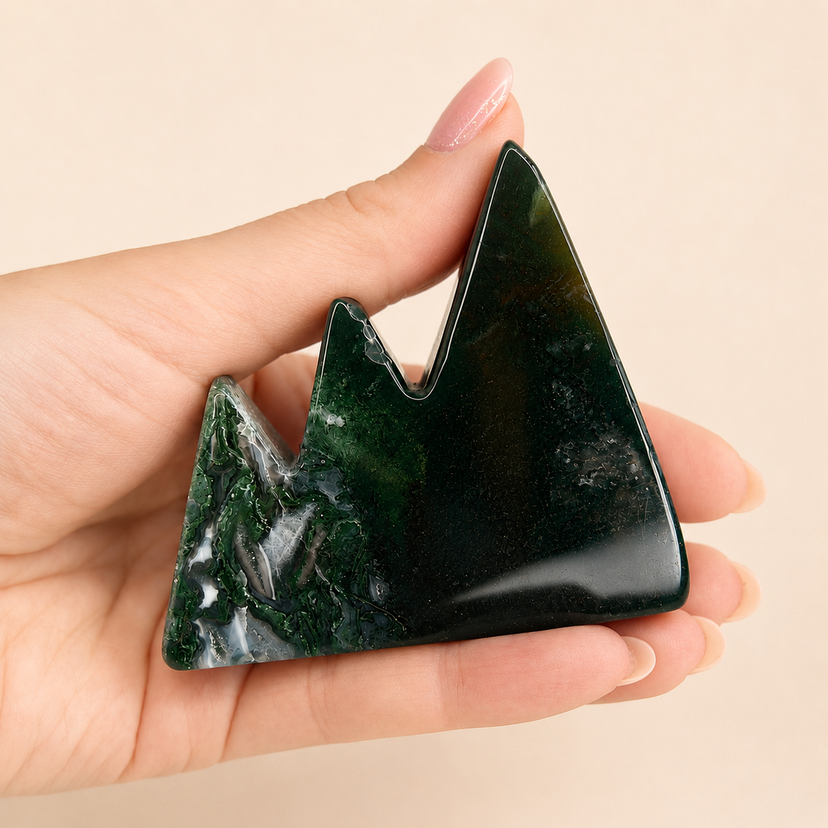 A hand holding a polished mountain-shaped dark green stone carving against a soft beige background.